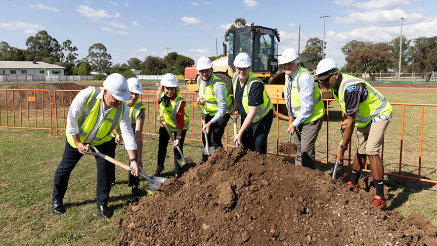 A collection of executive, board members and other staff in construction hats and vests with shovels performing the sod turn