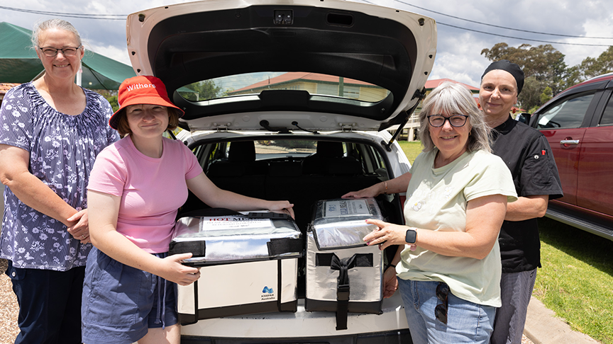 Three women stand beside a car, with a box open in the trunk, engaged in conversation and enjoying their time together.