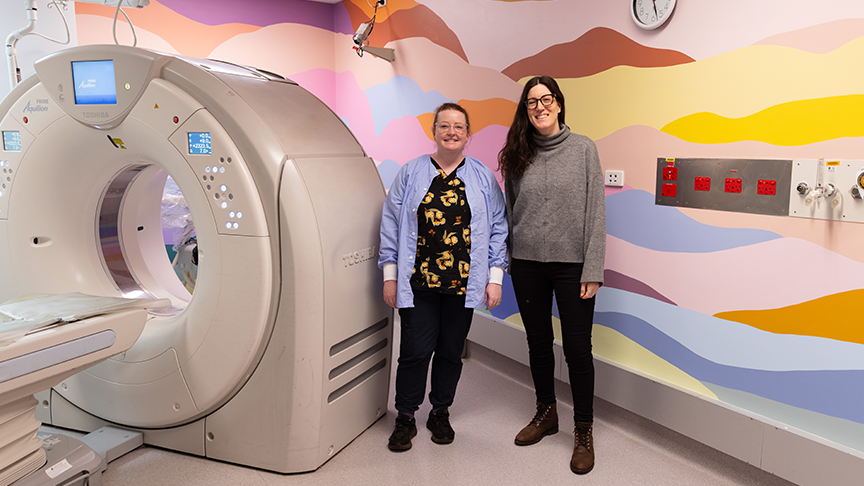 Two women standing in a room with a CT machine and colourful murals painted on the walls